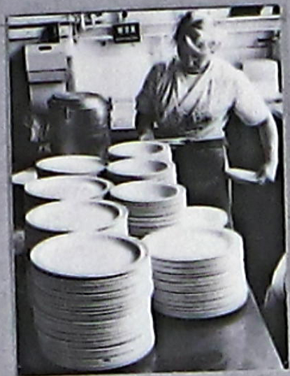 ‘Women and Work’ 1975 ©Hackney Flashers Image from ‘Women and Work’ 1975 shows a woman working in a kitchen with large stacks of plates in front of her.
