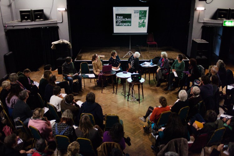 A view from above the audience. The people at the front including Julia Vellacott, Maggie Murray, Liz Heron and Sally Greenhill are turning to look behind them at a slide showing a poster for the exhibition "Three perspectives on photography"