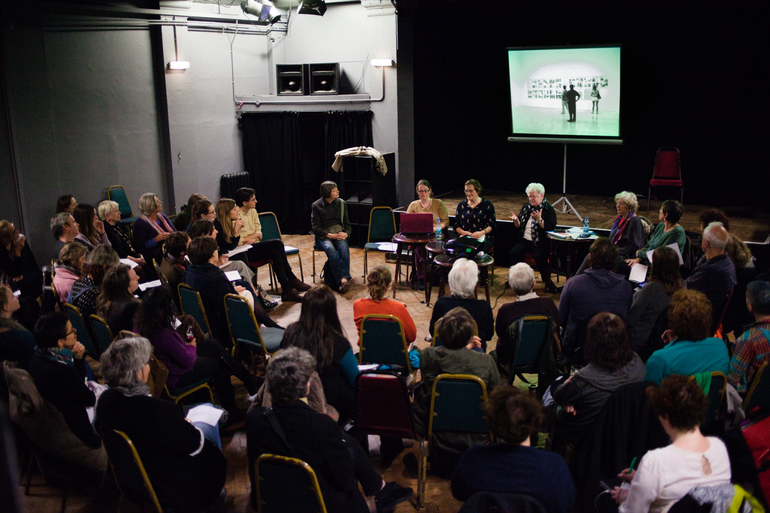 In a hall a large group of people seated in a semi-circle arrangement of chairs facing a panel listening to members of the Hackney Flashers speak about their work. In the centre of the front row is Maggie Murray, she is gesturing with her hands as she speaks. A projector behind shows an image of an exhibiton display and people viewing it.