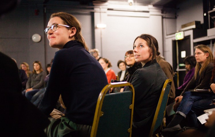 Two people seated in the audience turning to look towards the projector. Other people are seated around them.